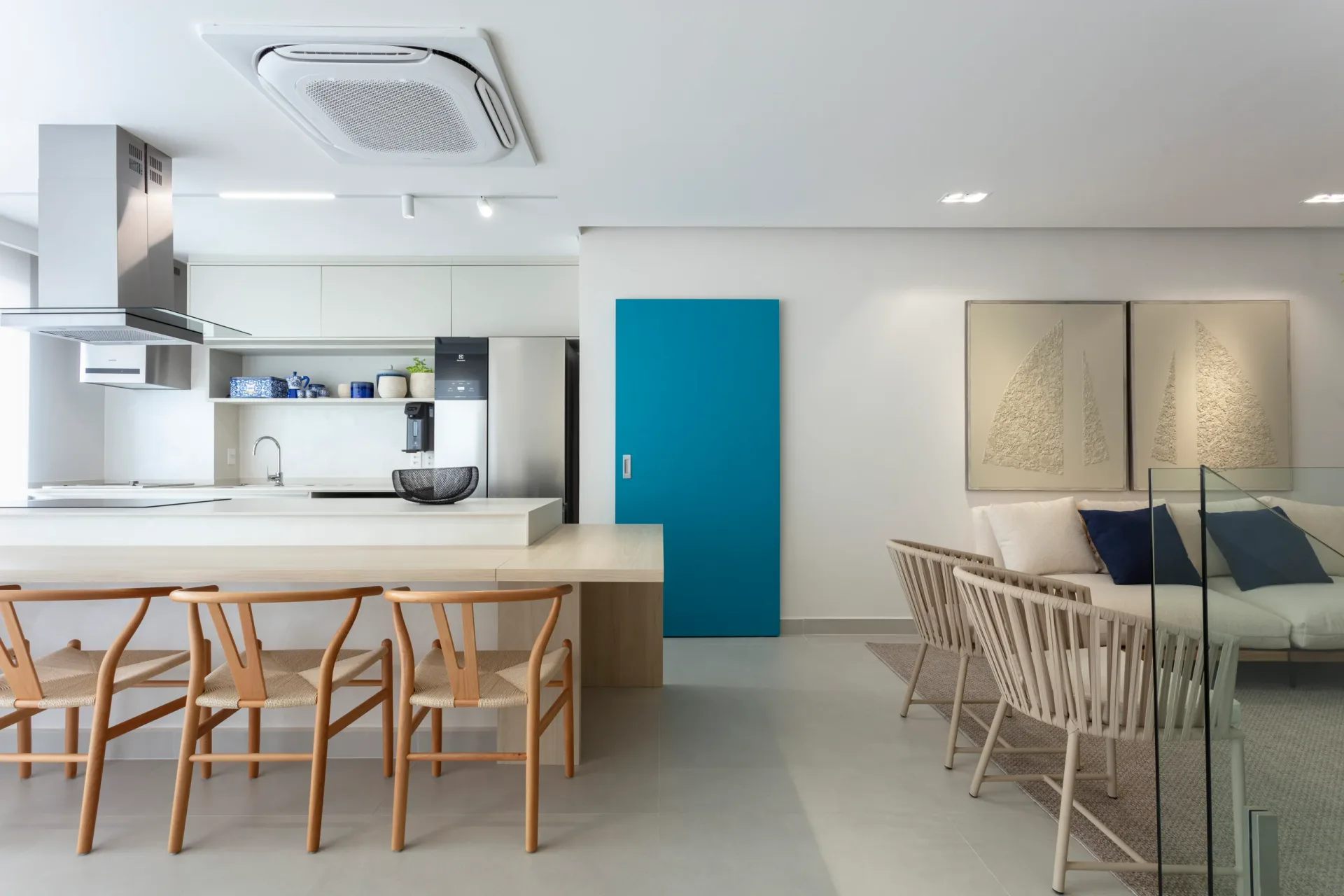 Cozinha branca integrada à sala de jantar, com bancada de madeira clara, banquetas e porta azul ao fundo